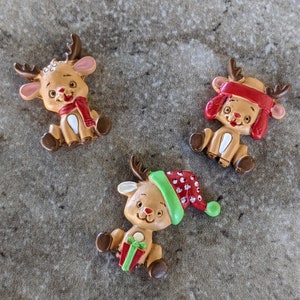 May include: Three small, brown reindeer figurines with painted details. One reindeer is wearing a red and white hat, another is wearing a green hat, and the third is wearing a red scarf.