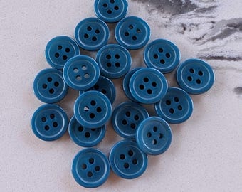 May include: A collection of teal-colored, round buttons with four holes each. The buttons are scattered on a light-colored surface. These buttons are suitable for sewing and craft projects.