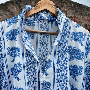 Può includere: Tunica bianca con stampa floreale blu, appesa a una gruccia di legno. Il design include fiori blu e strisce verticali. La parte superiore ha uno scollo a V ed è realizzata in tessuto leggero.