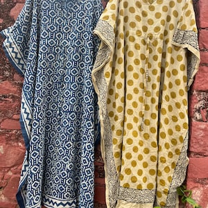 May include: Two kaftans hanging on light green plastic hangers. The left kaftan is navy blue with a white geometric pattern and a V-neckline. The right kaftan is beige with olive green polka dots, a V-neckline, and a black and white patterned border.
