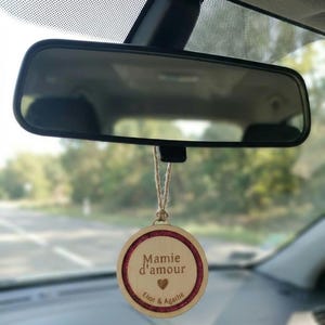 Puede incluir: Un adorno de coche de madera colgando del espejo retrovisor. El adorno redondo tiene un borde rojo brillante y las palabras "Mamie d'amour" y los nombres "Eliot & Agathe" están grabados. También hay un símbolo de corazón.