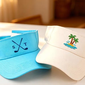 Könnte beinhalten: Ein hellblauer Sonnenschutz mit dunkelblau gestickten gekreuzten Golfschlägern und den Buchstaben S und T, neben einem weißen Sonnenschutz mit einer Stickerei von zwei Palmen auf einer kleinen Insel umgeben von blauen Wasserwellen.