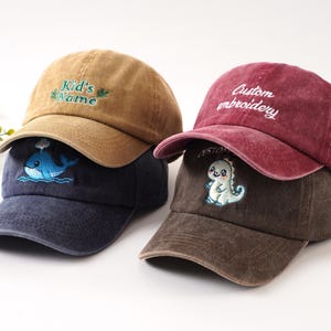 K&ouml;nnte beinhalten: Drei Baseballkappen in Braun-, Marine- und Burgundert&ouml;nen. Die braune Kappe hat den Text "Kid's Name" in Gr&uuml;n gestickt. Die marineblaue Kappe hat ein Wal-Design. Die bordeauxrote Kappe hat "Custom embroidery" in Wei&szlig;, und die braune Kappe hat einen Dinosaurier.
