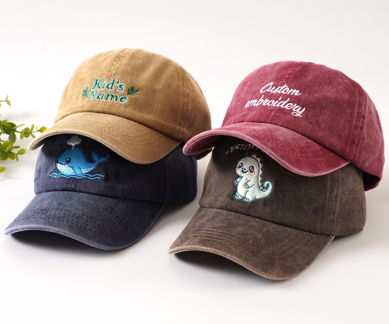 K&ouml;nnte beinhalten: Drei Baseballkappen in Braun-, Marine- und Burgundert&ouml;nen. Die braune Kappe hat den Text "Kid's Name" in Gr&uuml;n gestickt. Die marineblaue Kappe hat ein Wal-Design. Die bordeauxrote Kappe hat "Custom embroidery" in Wei&szlig;, und die braune Kappe hat einen Dinosaurier.