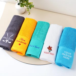 Puede incluir: Una colecci&oacute;n de seis toallas plegadas de colores. Cada toalla tiene un bordado diferente, incluyendo texto y dise&ntilde;os como "GOLF CLUB" y "Mom loves sports". Los colores incluyen gris, amarillo, turquesa, blanco y azul.