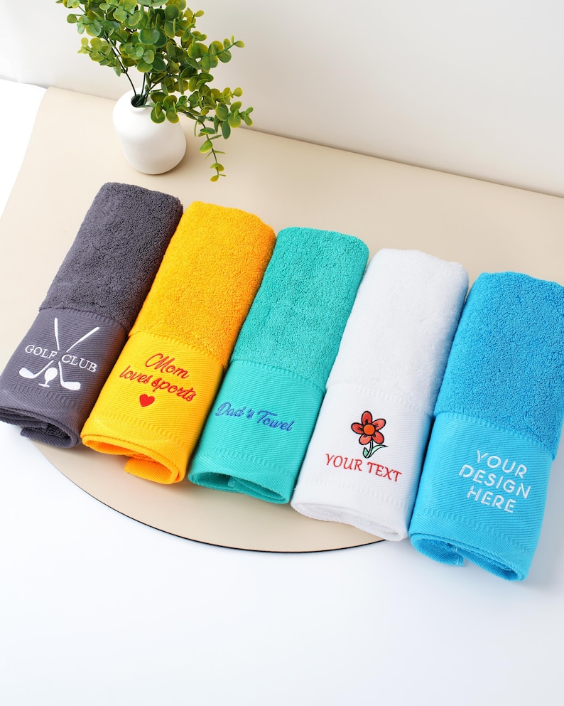 Puede incluir: Una colecci&oacute;n de seis toallas plegadas de colores. Cada toalla tiene un bordado diferente, incluyendo texto y dise&ntilde;os como "GOLF CLUB" y "Mom loves sports". Los colores incluyen gris, amarillo, turquesa, blanco y azul.