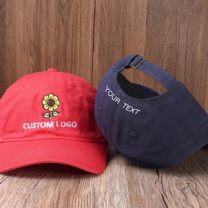 May include: Two baseball caps. One is red with a sunflower design and the text "CUSTOM LOGO". The other is navy blue with the text "YOUR TEXT" on the side. Both caps have adjustable straps.