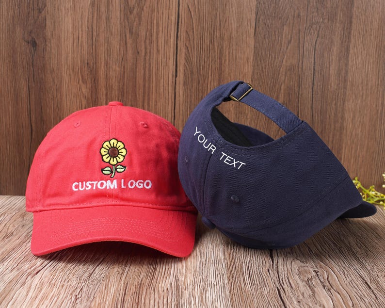 May include: Two baseball caps. One is red with a sunflower design and the text "CUSTOM LOGO". The other is navy blue with the text "YOUR TEXT" on the side. Both caps have adjustable straps.