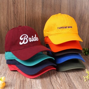 Pode incluir: Uma pilha de bonés de beisebol em várias cores, incluindo bordô, verde-azulado, azul marinho e vermelho. Um boné bordô apresenta a palavra "Bride" em escrita branca. Outro boné amarelo tem a frase "I LOVE MY MOM" em roxo.