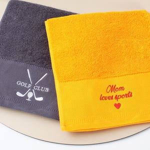 Puede incluir: Dos toallas de algod&oacute;n dobladas. Una es gris oscuro con "GOLF CLUB" bordado en blanco, con palos de golf cruzados y una pelota. La otra es amarilla con "Mom loves sports" y un coraz&oacute;n bordado en rojo.