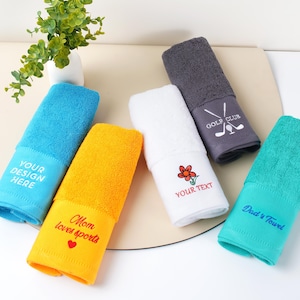 Puede incluir: Un conjunto de toallas enrolladas en colores vibrantes: azul, amarillo, blanco, gris y turquesa. Cada toalla tiene texto bordado, incluyendo "YOUR DESIGN HERE", "Mom loves sports", "YOUR TEXT", "Dad's Towel" y "GOLF CLUB".