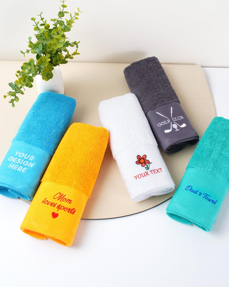 Puede incluir: Un conjunto de toallas enrolladas en colores vibrantes: azul, amarillo, blanco, gris y turquesa. Cada toalla tiene texto bordado, incluyendo "YOUR DESIGN HERE", "Mom loves sports", "YOUR TEXT", "Dad's Towel" y "GOLF CLUB".