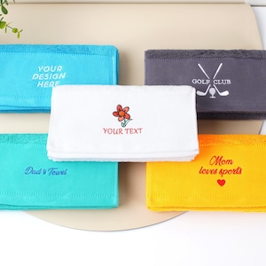 Puede incluir: Una colecci&oacute;n de toallas rectangulares plegadas en varios colores: azul, turquesa, blanco, gris y amarillo. Cada toalla presenta texto bordado o un gr&aacute;fico, incluyendo "YOUR DESIGN HERE", "Dad's Towel", una flor, "GOLF CLUB" y "Mom loves sports".