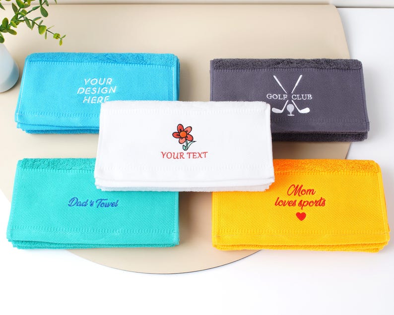 Puede incluir: Una colecci&oacute;n de toallas rectangulares plegadas en varios colores: azul, turquesa, blanco, gris y amarillo. Cada toalla presenta texto bordado o un gr&aacute;fico, incluyendo "YOUR DESIGN HERE", "Dad's Towel", una flor, "GOLF CLUB" y "Mom loves sports".