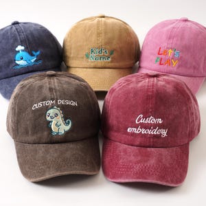 K&ouml;nnte beinhalten: F&uuml;nf Baseballkappen in verschiedenen Farben: Marineblau mit Wal, Beige mit "Kid's Name", Rosa mit "Let's PLAY", Braun mit Dinosaurier und Burgunderrot mit "Custom embroidery". Alle Kappen haben einen gebogenen Schirm.