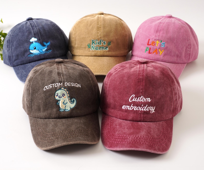 K&ouml;nnte beinhalten: F&uuml;nf Baseballkappen in verschiedenen Farben: Marineblau mit Wal, Beige mit "Kid's Name", Rosa mit "Let's PLAY", Braun mit Dinosaurier und Burgunderrot mit "Custom embroidery". Alle Kappen haben einen gebogenen Schirm.
