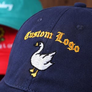 May include: Navy blue baseball cap with "Custom Logo" embroidered in gold lettering. A white goose with yellow feet is embroidered below the text. A red cap with a sunflower design and a green cap are visible in the background.