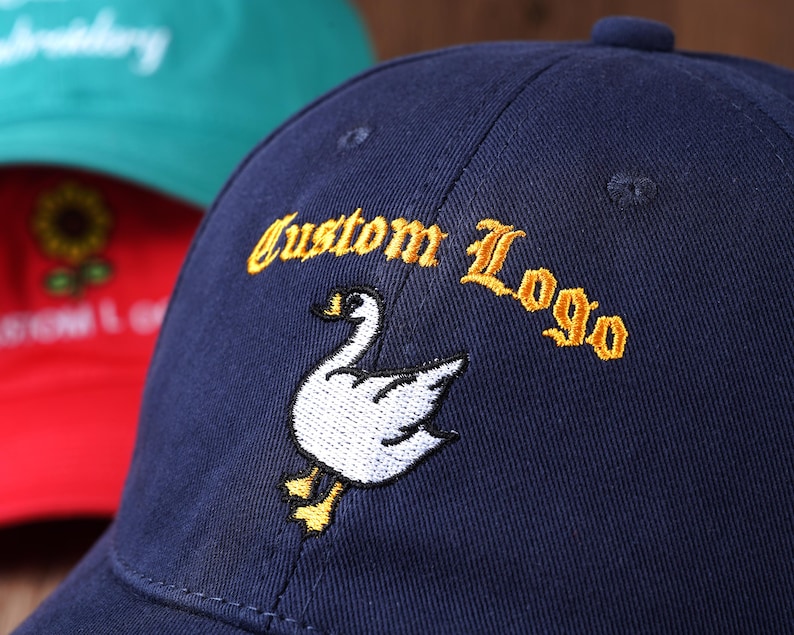 May include: Navy blue baseball cap with "Custom Logo" embroidered in gold lettering. A white goose with yellow feet is embroidered below the text. A red cap with a sunflower design and a green cap are visible in the background.