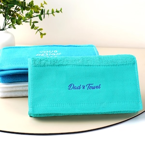 Puede incluir: Toallas dobladas en tonos azules y blancos. La toalla delantera es de color turquesa con el texto bordado "Dad's Towel" en azul. Otra toalla en el fondo tiene el texto "YOUR DESIGN".