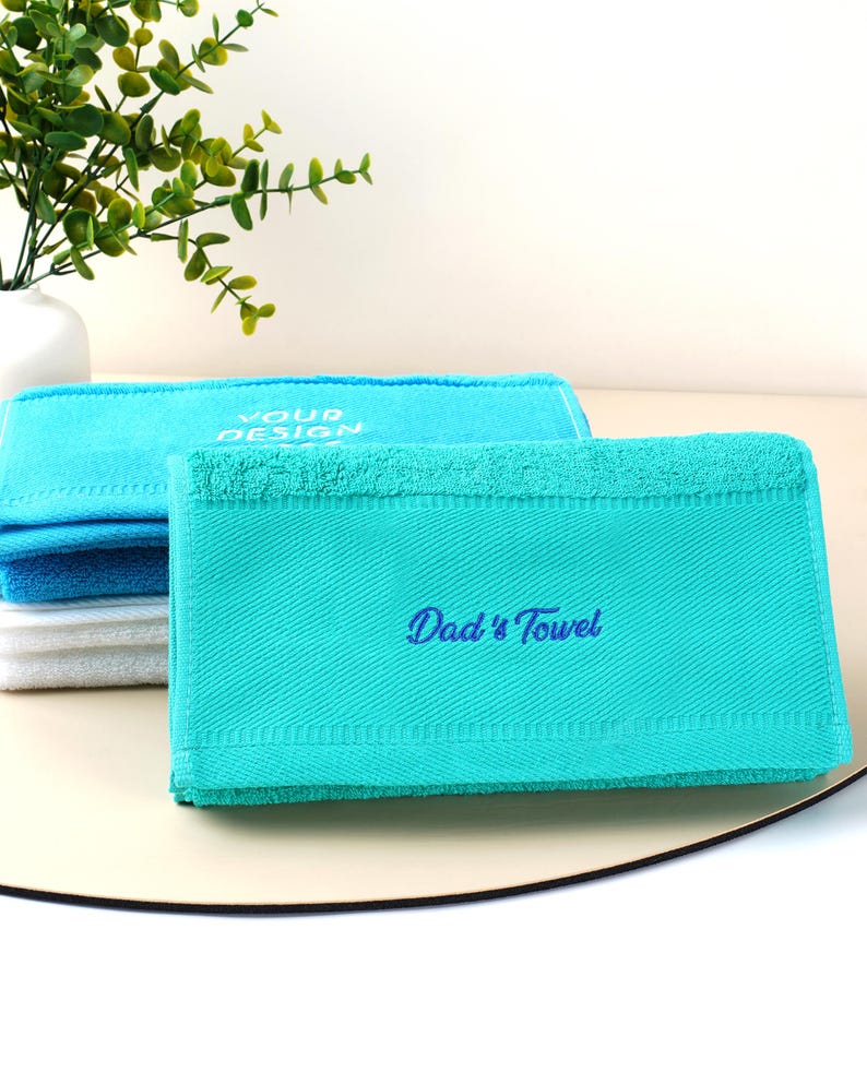 Puede incluir: Toallas dobladas en tonos azules y blancos. La toalla delantera es de color turquesa con el texto bordado "Dad's Towel" en azul. Otra toalla en el fondo tiene el texto "YOUR DESIGN".