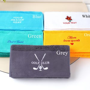 Puede incluir: Una pila de toallas plegadas en varios colores: azul, verde, blanco, naranja y gris. Cada toalla tiene texto bordado o un dise&ntilde;o, incluyendo "YOUR DESIGN HERE", "Dad's Towel", una flor, "YOUR TEXT", "Mom loves sports" y "GOLF CLUB".