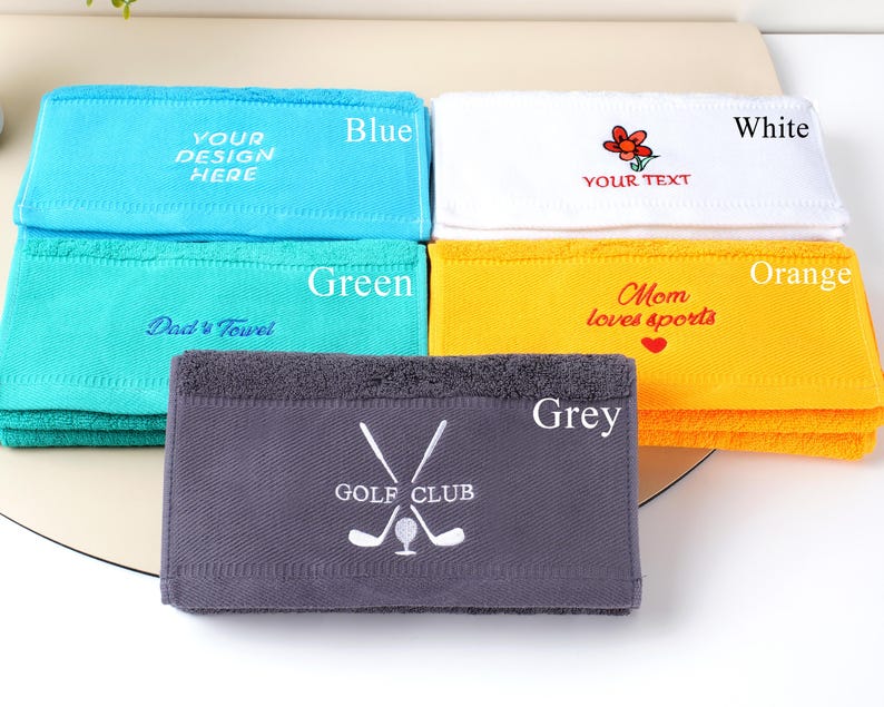 Puede incluir: Una pila de toallas plegadas en varios colores: azul, verde, blanco, naranja y gris. Cada toalla tiene texto bordado o un dise&ntilde;o, incluyendo "YOUR DESIGN HERE", "Dad's Towel", una flor, "YOUR TEXT", "Mom loves sports" y "GOLF CLUB".