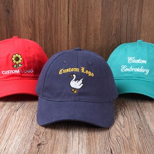 Può includere: Tre cappellini da baseball rossi, blu navy e verde acqua. Il cappellino rosso presenta un girasole e la scritta "CUSTOM LOGO". Il cappellino blu navy ha una grafica di un'oca e "Custom Logo". Il cappellino verde acqua mostra "Custom Embroidery".