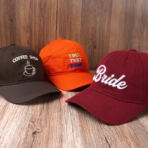 May include: Three baseball caps in a row. One is dark brown with "Coffee Shop" embroidered in white, another is orange with "YOUR TEXT HERE" in colorful thread, and the last is burgundy with "Bride" in white.