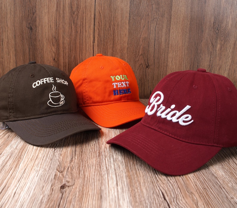 May include: Three baseball caps in a row. One is dark brown with "Coffee Shop" embroidered in white, another is orange with "YOUR TEXT HERE" in colorful thread, and the last is burgundy with "Bride" in white.