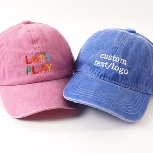 K&ouml;nnte beinhalten: Zwei Baseballkappen, eine rosa und eine blaue. Die rosa Kappe hat den gestickten Text "Let's PLAY" in bunten F&auml;den. Die blaue Kappe zeigt den gestickten Text "custom text/logo" in Wei&szlig;. Beide Kappen haben einen gebogenen Schirm.
