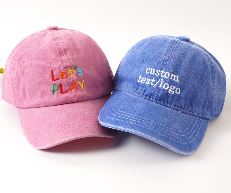 K&ouml;nnte beinhalten: Zwei Baseballkappen, eine rosa und eine blaue. Die rosa Kappe hat den gestickten Text "Let's PLAY" in bunten F&auml;den. Die blaue Kappe zeigt den gestickten Text "custom text/logo" in Wei&szlig;. Beide Kappen haben einen gebogenen Schirm.