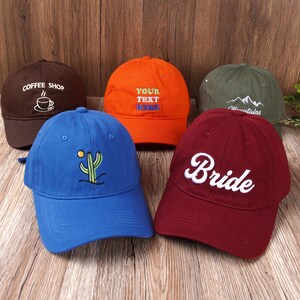 May include: A collection of baseball caps in various colors, including brown, orange, blue, green, and burgundy. Each cap features unique embroidered designs or text, such as a coffee cup, cactus, mountains, and the word "Bride".