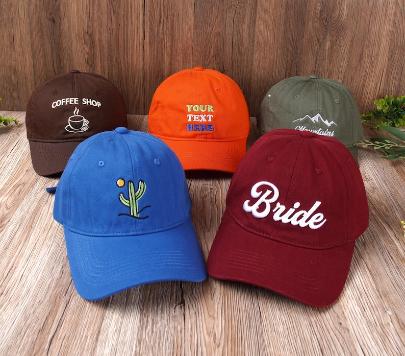 May include: A collection of baseball caps in various colors, including brown, orange, blue, green, and burgundy. Each cap features unique embroidered designs or text, such as a coffee cup, cactus, mountains, and the word "Bride".