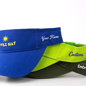 May include: Three sun visors stacked together. The top visor is royal blue with a yellow sun icon and the text COOLE HAT. The middle visor is lime green with the text Your Name. The bottom visor is dark olive green with the text Embroidery.