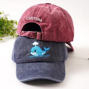 K&ouml;nnte beinhalten: Zwei Baseballkappen. Die obere Kappe ist tiefrot mit "CUSTOM" auf der R&uuml;ckseite gestickt. Die untere Kappe ist verwaschen blau mit einem gestickten blauen Wal-Design. Beide Kappen haben verstellbare Metallschnallen.