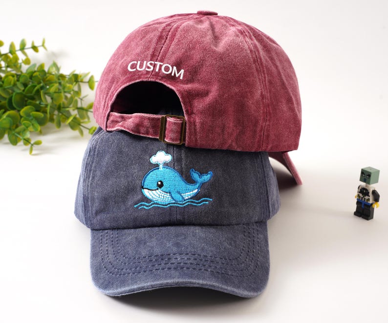 K&ouml;nnte beinhalten: Zwei Baseballkappen. Die obere Kappe ist tiefrot mit "CUSTOM" auf der R&uuml;ckseite gestickt. Die untere Kappe ist verwaschen blau mit einem gestickten blauen Wal-Design. Beide Kappen haben verstellbare Metallschnallen.