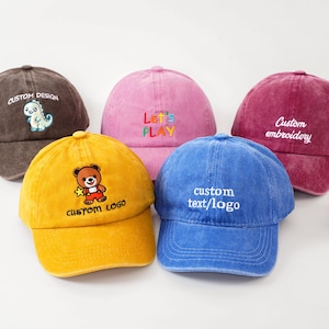 Puede incluir: Cinco gorras de béisbol coloridas en marrón, rosa, amarillo, azul y granate. Cada gorra presenta diseños y texto bordados, incluyendo un dinosaurio, un oso y las palabras "Let's PLAY" y "Custom text/logo".