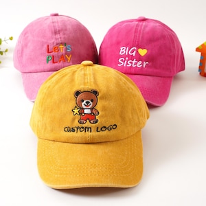 K&ouml;nnte beinhalten: Drei Baseballkappen f&uuml;r Kinder. Eine gelbe Kappe mit einem B&auml;ren und dem Text "CUSTOM LOGO". Eine rosa Kappe mit "BIG Sister" gestickt. Eine weitere rosa Kappe mit "Let's PLAY".