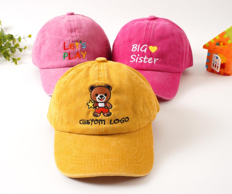 K&ouml;nnte beinhalten: Drei Baseballkappen f&uuml;r Kinder. Eine gelbe Kappe mit einem B&auml;ren und dem Text "CUSTOM LOGO". Eine rosa Kappe mit "BIG Sister" gestickt. Eine weitere rosa Kappe mit "Let's PLAY".