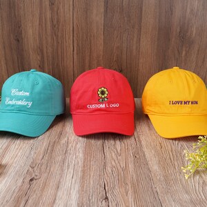 May include: Three baseball caps in teal, red, and yellow. The teal cap has "Custom Embroidery" embroidered in white. The red cap features a sunflower design and "CUSTOM LOGO". The yellow cap says "I LOVE MY MOM".