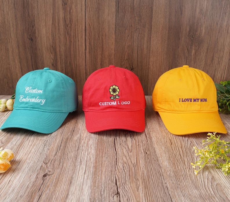 May include: Three baseball caps in teal, red, and yellow. The teal cap has "Custom Embroidery" embroidered in white. The red cap features a sunflower design and "CUSTOM LOGO". The yellow cap says "I LOVE MY MOM".