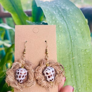 Op de afbeelding: Handgemaakte oorbellen met een schelpenhanger met bruine en witte vlekken, gezet in een geweven, cirkelvormig frame. De oorbellen hebben goudkleurige haken en worden gepresenteerd op een bruine kaart.