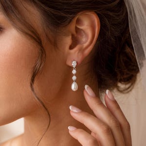 Può includere: Un orecchino pendente color argento con una pietra trasparente a forma di pera in alto, seguita da due pietre rotonde trasparenti e terminata con una perla bianca a forma di goccia nella parte inferiore. Fissaggio a perno.