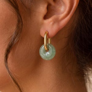 Puede incluir: Un pendiente de aro de metal dorado con un colgante de piedra circular de color verde pálido. El aro de oro pulido tiene una forma ovalada alargada y pasa a través del orificio central de la piedra translúcida de color verde claro.