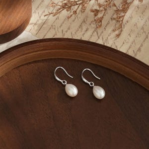 Pode incluir: Um par de brincos pendentes com pérolas brancas em forma de gota, suspensas por ganchos simples de metal prateado. Os brincos possuem um acabamento liso e brilhante, destacando a forma natural das pérolas em seu design.