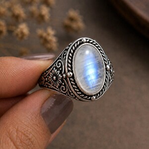 May include: A silver ring featuring an oval, iridescent moonstone. The ring has an ornate, floral design on the band and around the stone. The moonstone displays a blue and white color. The ring is a piece of jewelry.