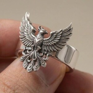 May include: A silver ring featuring a detailed, three-dimensional phoenix with wings spread wide and a long, ornate tail. The metal has a polished finish on the band and a darkened, oxidized finish on the bird to highlight the intricate feather textures.