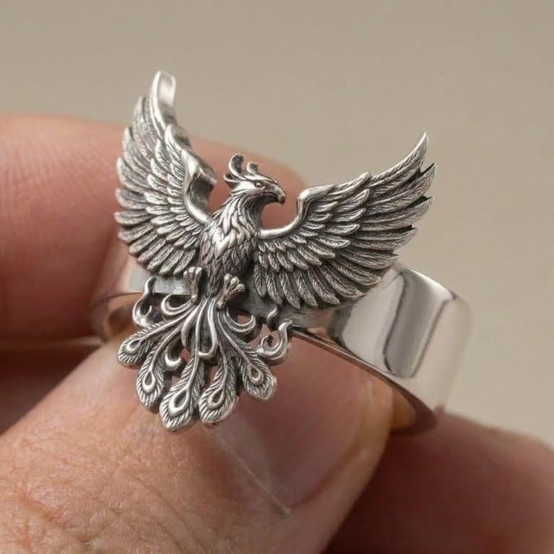 May include: A silver ring featuring a detailed, three-dimensional phoenix with wings spread wide and a long, ornate tail. The metal has a polished finish on the band and a darkened, oxidized finish on the bird to highlight the intricate feather textures.