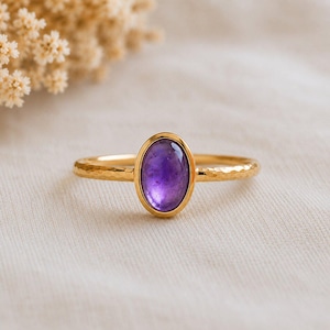 Può includere: Un anello d'oro con una pietra preziosa ovale viola. L'anello ha una texture martellata sulla fascia e un castone liscio e lucido. La gemma è di un colore viola intenso. L'anello è posto su uno sfondo neutro.