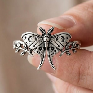 Puede incluir: Un anillo de tono plateado con una polilla detallada en el centro, decorada con motivos grabados de lunas crecientes y estrellas en las alas. La banda del anillo se extiende en delicadas ramas con hojas a ambos lados del diseño central.